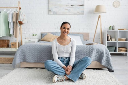Téléchargez les photos : Étudiant afro-américain joyeux en jeans assis avec les jambes croisées près du lit à la maison - en image libre de droit
