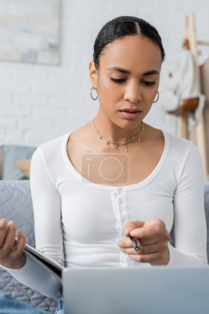 estudiante afroamericano inteligente sosteniendo el cuaderno y señalando con la pluma en el ordenador portátil 