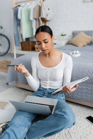 Foto de Estudiante afroamericano inteligente sosteniendo el cuaderno y la pluma mientras que mira webinar en computadora portátil - Imagen libre de derechos