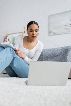hübsche afrikanisch-amerikanische Frau in Jeans macht sich Notizen, während sie Online-Vorlesung auf Laptop anschaut 