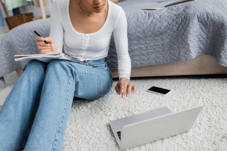abgeschnittene Ansicht einer afrikanisch-amerikanischen Frau in Jeans, die Notizen macht, während sie Online-Vorlesung am Laptop anschaut 