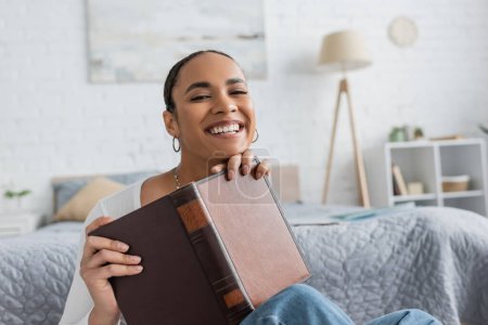 pleased african american student holding book while studying from home 