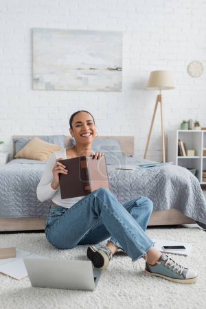Photo for Pleased african american student holding book while sitting near laptop and studying from home - Royalty Free Image