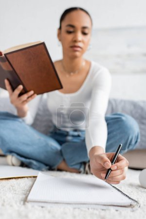 Foto de Vista de ángulo bajo del joven estudiante afroamericano sentado con libro mientras toma notas en el cuaderno - Imagen libre de derechos