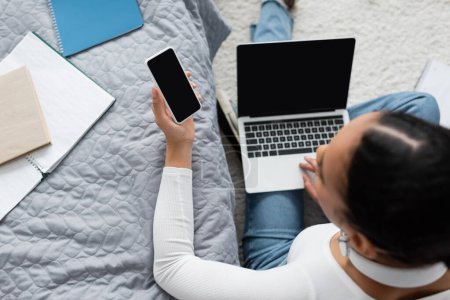 Téléchargez les photos : Vue du haut de la femme afro-américaine tenant smartphone avec écran vide près d'un ordinateur portable tout en étudiant de la maison - en image libre de droit