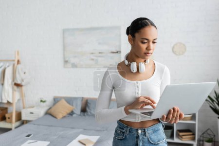 Foto de Estudiante afroamericano bonito con auriculares inalámbricos en el cuello usando el ordenador portátil en dormitorio moderno - Imagen libre de derechos