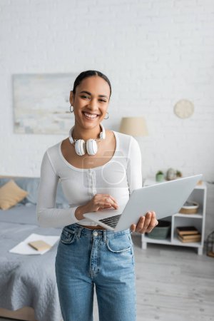 Foto de Estudiante afroamericano feliz con auriculares inalámbricos en el cuello usando el ordenador portátil en el dormitorio moderno - Imagen libre de derechos