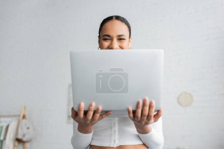 Foto de Estudiante afroamericano feliz cubriendo la mitad de la cara mientras sostiene el ordenador portátil - Imagen libre de derechos