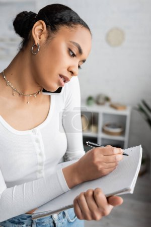 Foto de Joven estudiante afroamericano hablando en un teléfono inteligente mientras escribe en un cuaderno - Imagen libre de derechos
