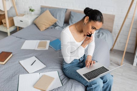 Photo for Overhead view of african american student talking on smartphone while using laptop and studying online from home - Royalty Free Image