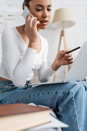 Foto de Joven afroamericano estudiante hablando en el teléfono inteligente mientras mira el ordenador portátil y aprender en línea desde casa - Imagen libre de derechos
