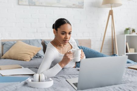 junge afrikanisch-amerikanische Studentin hält Tasse Kaffee in der Hand und schaut auf Laptop im modernen Schlafzimmer 