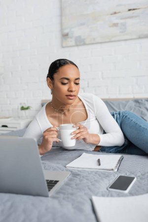 Foto de Joven afroamericano estudiante sosteniendo taza de café y mirando portátil cerca de portátil en dormitorio moderno - Imagen libre de derechos