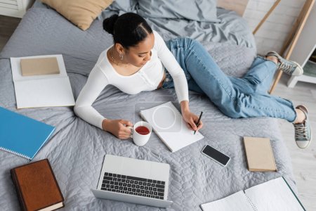 Draufsicht auf afrikanisch-amerikanische Studentin, die eine Tasse Tee hält und im Schlafzimmer auf Notizbuch schreibt 
