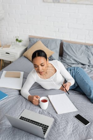 Afrikanischer Student hält Tasse Tee in der Nähe von Laptop und Notizbuch auf dem Bett zu Hause 