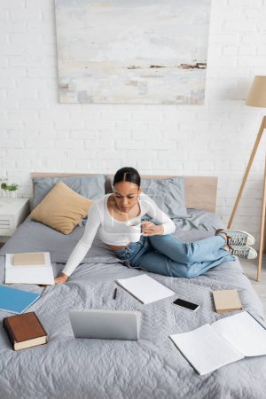 Estudiante afroamericano sosteniendo taza cerca de dispositivos y cuadernos en la cama en casa 