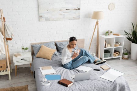 Estudiante afroamericano sosteniendo taza de té cerca de gadgets y libros en la cama en casa 