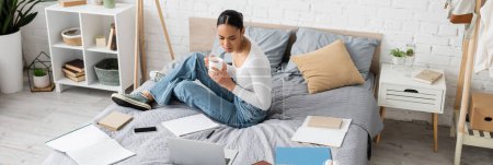 African american student holding tea near devices and notebooks on bed in bedroom, banner 