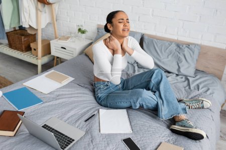 Exhausted african american student touching neck near devices and notebooks on bed at home 