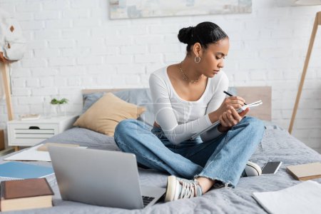 African american student writing on notebook near devices on bed at home 