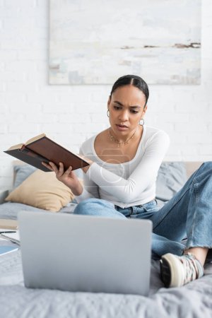 Konzentrierte afrikanisch-amerikanische Studentin hält Buch in der Hand und schaut zu Hause auf Laptop im Bett 