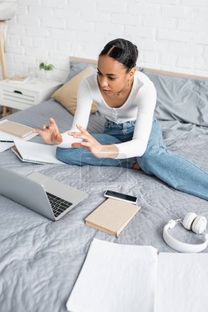 African american student talking during webinar on laptop near books on bed at home 