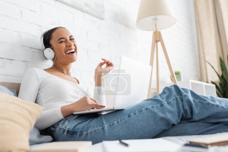 Alegre estudiante afroamericano en auriculares usando computadora portátil en la cama en casa 