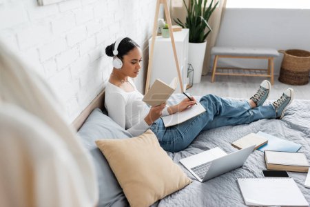 African american student in headphones reading book and writing on notebook near devices on bed 