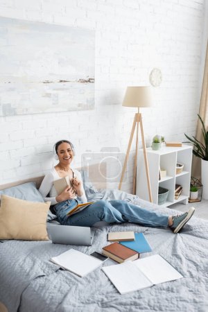 Cheerful african american student in headphones holding book near gadgets on bed 