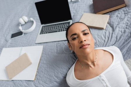 Top view of african american student looking at camera near devices and books on bed 