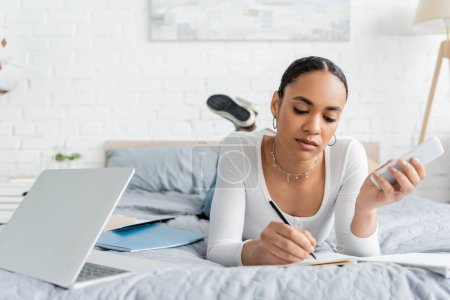 African american student holding smartphone and writing on notebook near laptop on bed 