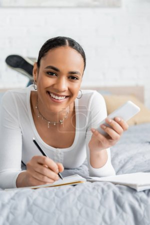 Positive african american student holding cellphone and writing on notebook on bed 