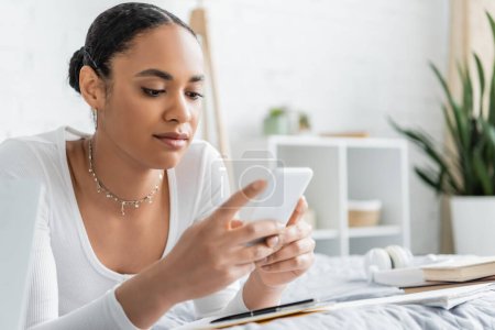 Smiling african american student using smartphone near blurred books on bed 