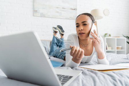 African american student talking on smartphone and looking at blurred laptop while lying on bed 