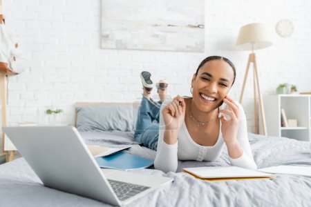 Smiling african american student talking on smartphone near notebooks and laptop in bedroom 