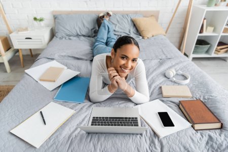 Happy african american student lying near devices and books on bed 