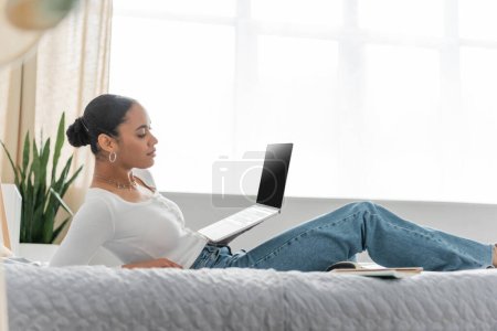 African american student using laptop with blank screen on bed at home 