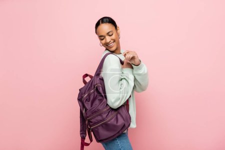 Bonita y sonriente estudiante afroamericana mirando mochila aislada en rosa 