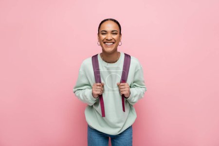 Foto de Sonriente estudiante afroamericano sosteniendo mochila aislada en rosa - Imagen libre de derechos
