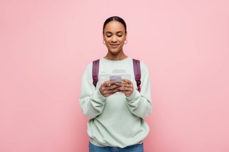 Smiling african american student with backpack typing message on smartphone isolated on pink 