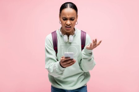 Étudiant afro-américain en colère avec écouteurs utilisant un téléphone mobile isolé sur rose 