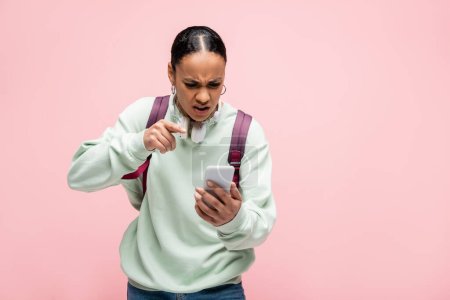 Étudiant afro-américain en colère avec sac à dos et écouteurs pointant vers smartphone isolé sur rose 