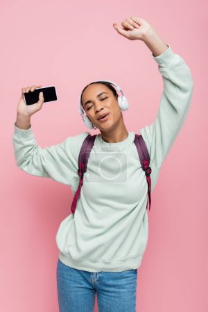 young african american student in wireless headphones holding smartphone while listening music and singing on pink  Poster 647409974