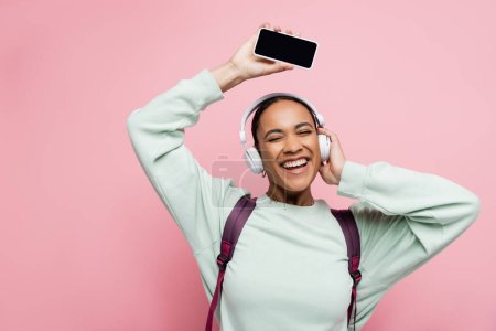 Happy african american woman in headphones holding smartphone with blank screen and listening music on pink background