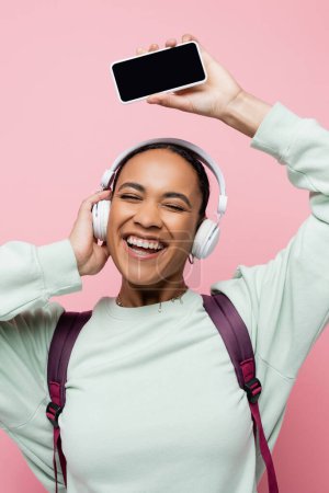 Overjoyed african american woman in wireless headphones holding smartphone while listening music isolated on pink