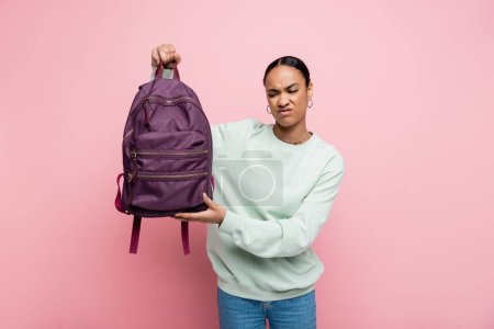 displeased african american student in sweatshirt holding purple backpack isolated on pink 