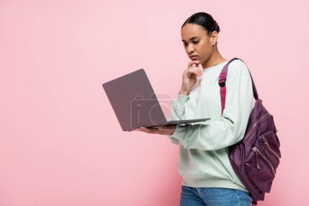 estudiante afroamericano pensativo con la mochila usando el ordenador portátil en el fondo rosa 