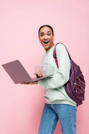 erstaunt afrikanisch-amerikanischer Student mit Rucksack, Laptop in der Hand und Blick auf Kamera isoliert auf rosa 