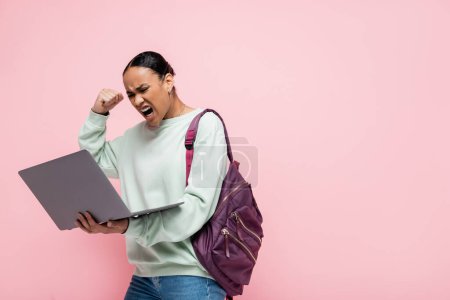 Irritierte afrikanisch-amerikanische Studentin mit Rucksack blickt auf Laptop isoliert auf rosa 