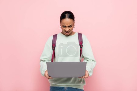 estudiante afroamericano enojado con la mochila usando el ordenador portátil aislado en rosa 